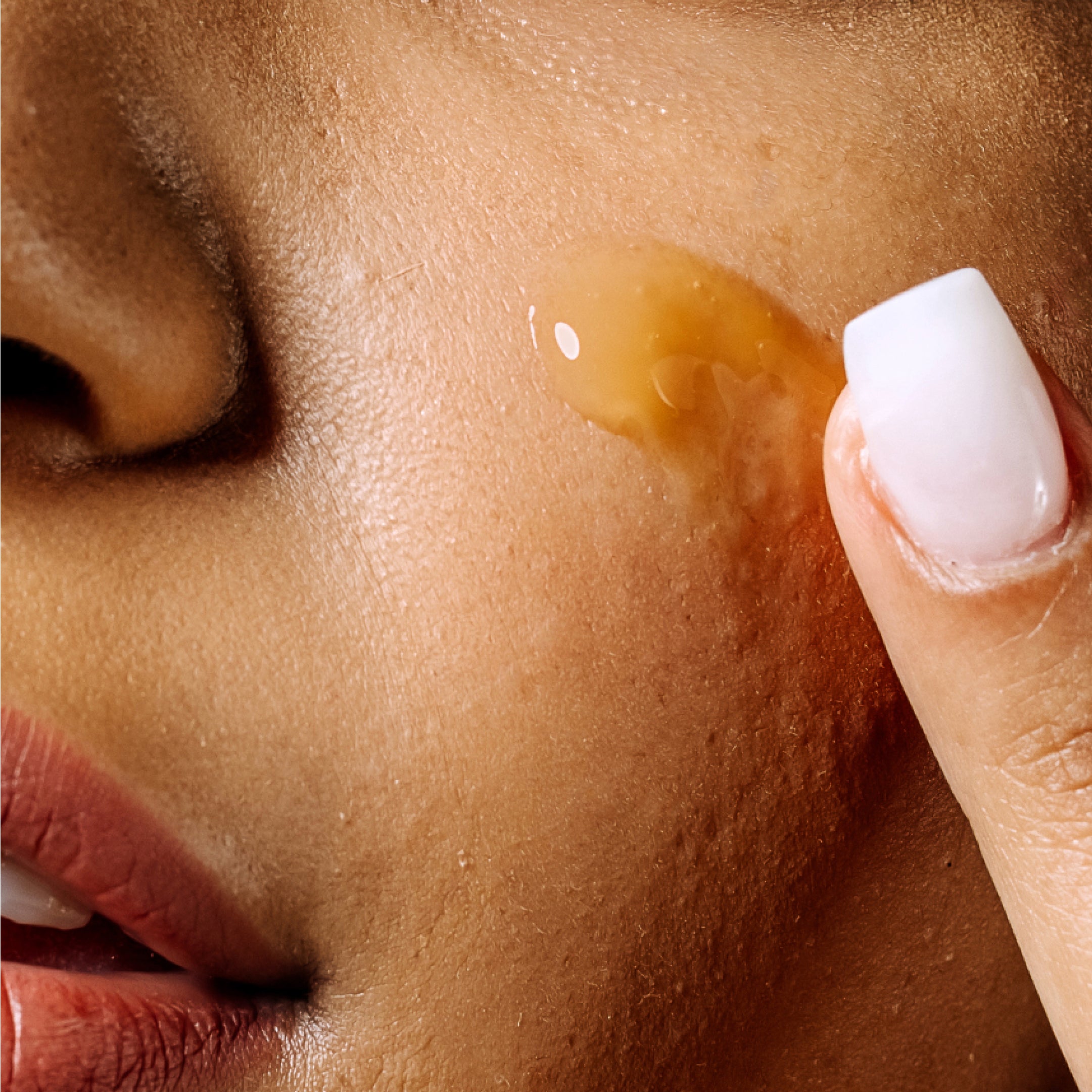 Close-up of a person applying Geologie's Vitamin C+E Ferulic Serum to cheek, focusing on skin and fingertips with a droplet of serum visible.